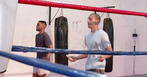 Boxing ring duo skipping rope while training partner warming up by heavy bags in gym