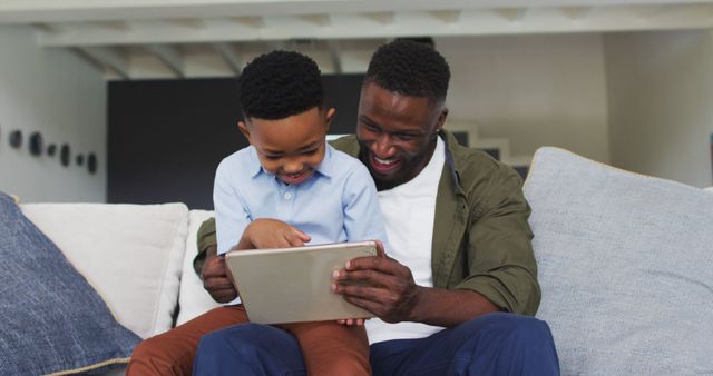 Father and son sitting on a couch in a cozy living room, engaging with a digital tablet, both smiling and enjoying their time together. This can be used for advertisements about family products, technology in family life, or creating memorable moments at home.