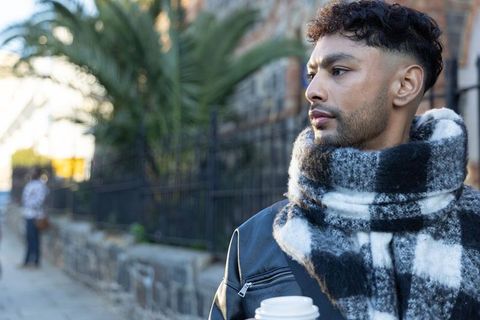 Man holding takeaway coffee wearing checkered scarf and leather jacket on city sidewalk