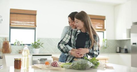 Sunlit same-sex female couple embracing at kitchen island preparing fresh produce