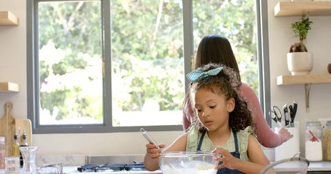 Mother and daughter baking together in sunlit kitchen stirring batter with whisk