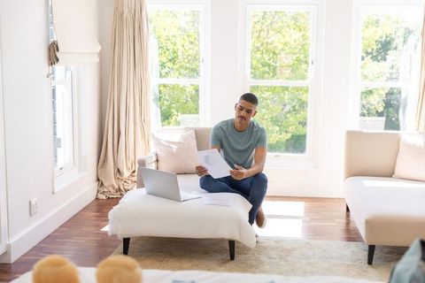 Man reviewing documents in sunlit living room while working on laptop for remote work