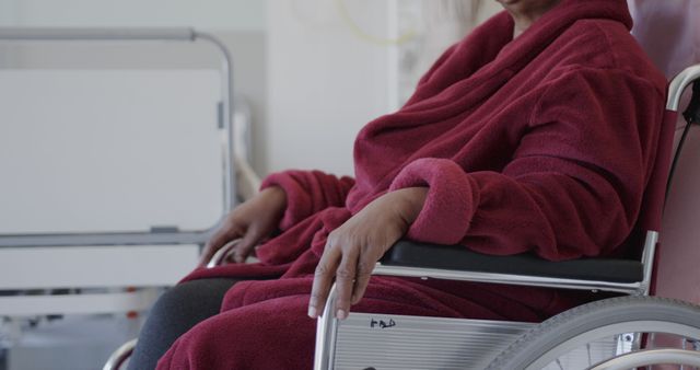 Elderly woman in a red robe sitting in a wheelchair at a hospital room. She is partially visible, with focus on her hands and robe. Ideal for healthcare promotions, senior care advertisements, and medical service brochures.