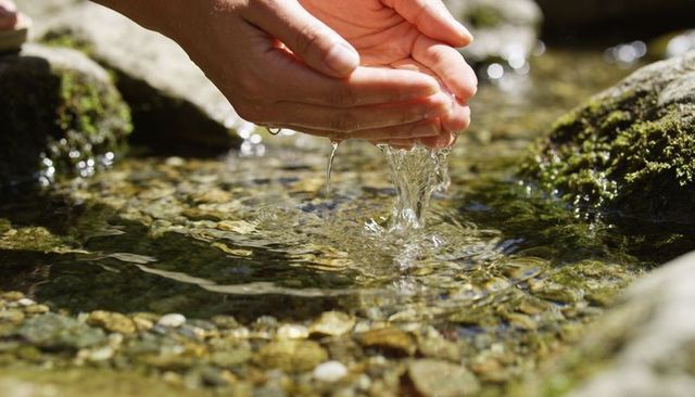Cupped Hands Collecting Clear Creek Water Over Pebbled Stream with Sunlit Ripples