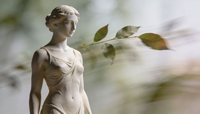 Classical female sculpture with flowing drapery and motion-blur leaves in soft garden light
