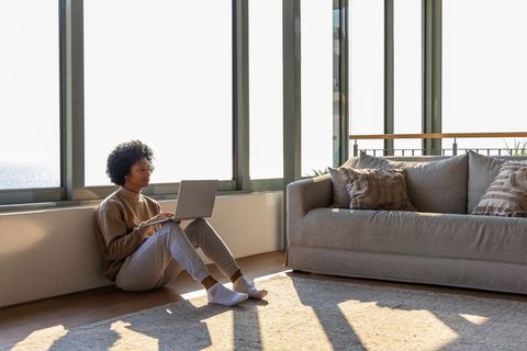 African american woman working on laptop by sunlit windows in minimalist living room
