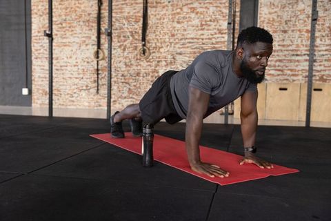 African american man performing plank on red mat in industrial gym with rings