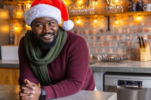 African american man wearing santa hat and scarf leaning on counter smiling in rustic kitchen