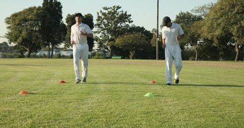 Diverse male cricket teammates jogging between cones on grass field for outdoor training