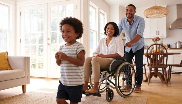 Father pushing partner in wheelchair while child runs through sunlit modern family home