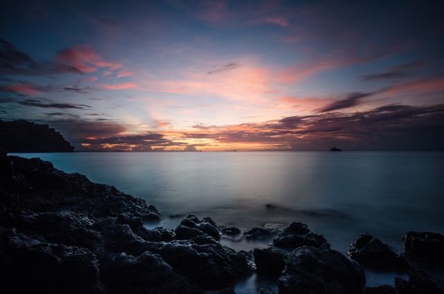Sunset over ocean where horizon and sea blend seamlessly. Dark rocks in foreground. Uses: travel inspiration, website backgrounds, relaxation themes, nature-related content, scenic calendars.