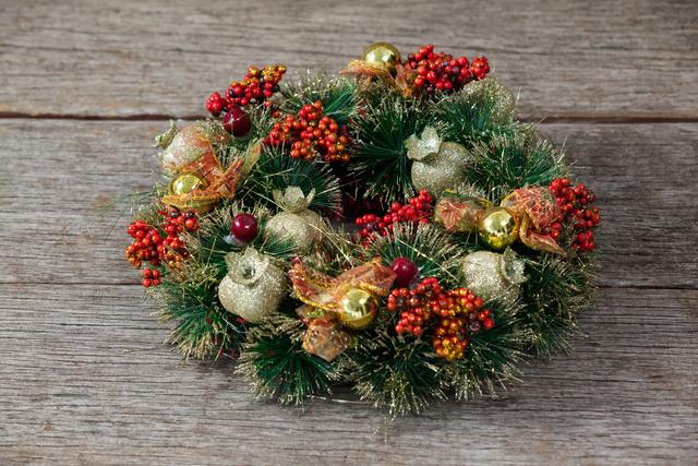 Close-up of a beautifully decorated Christmas wreath placed on a rustic wooden plank. The wreath features green pine branches, red berries, gold ornaments, and festive ribbons. Ideal for use in holiday-themed designs, greeting cards, seasonal promotions, and festive home decor inspiration.