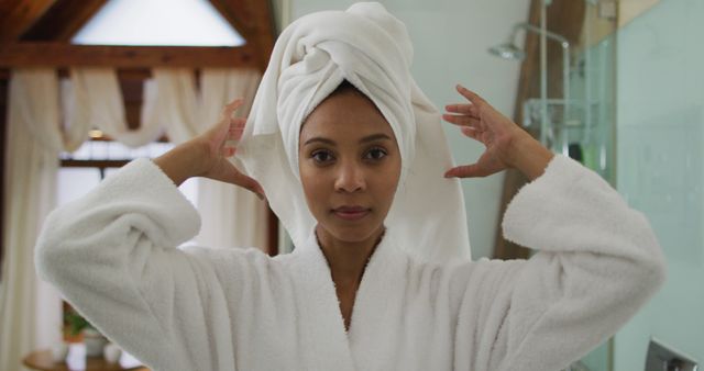 A woman wearing a bathrobe is adjusting a towel wrapped around her wet hair in a bright bathroom. This can be used for topics related to self-care, daily routines, hygiene, and relaxation products. Perfect for spa, wellness, grooming, and beauty industry promotions.