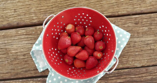 A red colander filled with fresh strawberries sits on a wooden surface, with copy space. Ripe berries signify the start of summer and evoke thoughts of healthy eating and seasonal cooking.