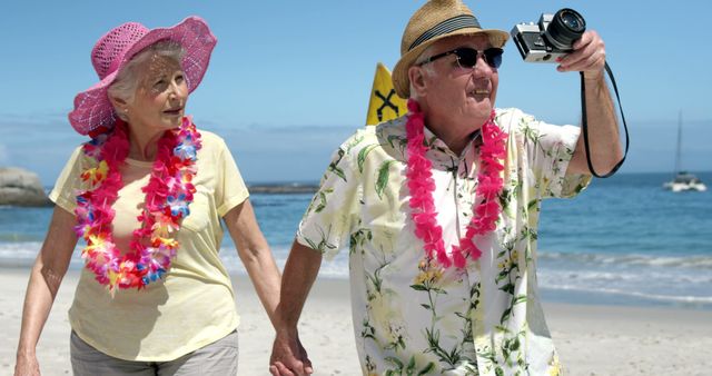 Senior couple enjoying vacation, walking on beach with Hawaiian leis, holding hands while taking photos. Perfect for concepts of retirement, leisure, travel, and togetherness in senior years.