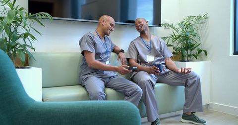 African american healthcare workers laughing in clinic break room holding phone and mug