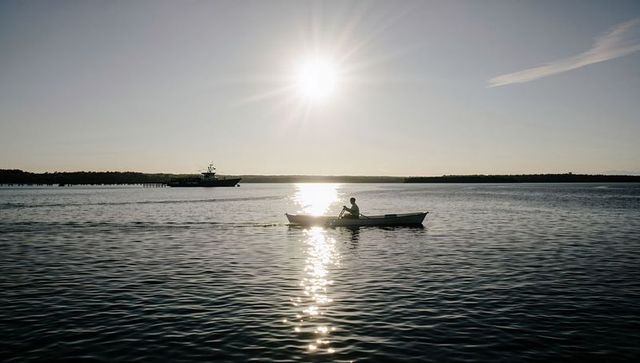 Solo rower paddling through sun glare over calm water with horizon boat silhouette