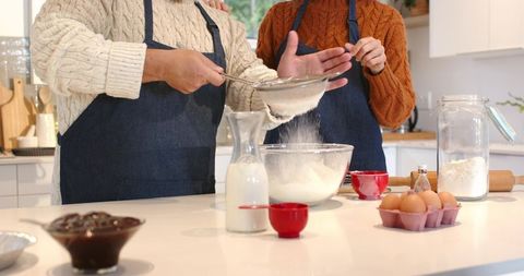Couple sifting flour together in sunlit kitchen wearing denim aprons for baking