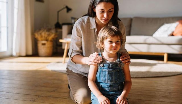 Mother kneeling with toddler in denim overalls bonding by sunlight in modern living room