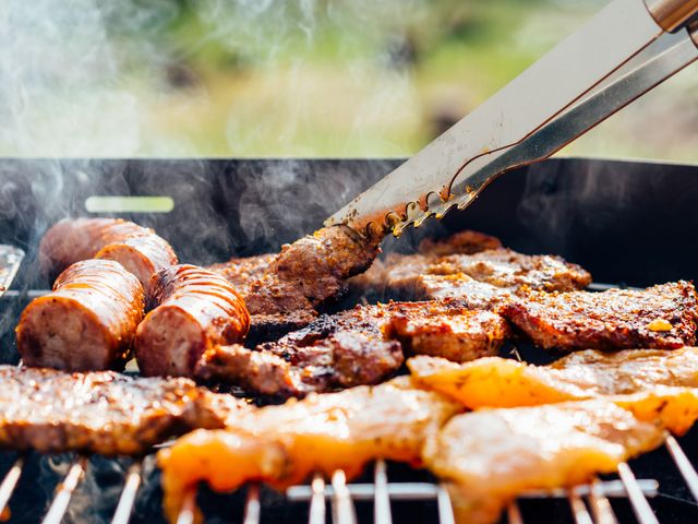 Close-up of various meats including sausages and steaks grilling on a barbecue with smoke rising and tongs turning one piece. Perfect depiction for summer cooking, outdoor gatherings, BBQ recipes, and food-related advertisements or articles.