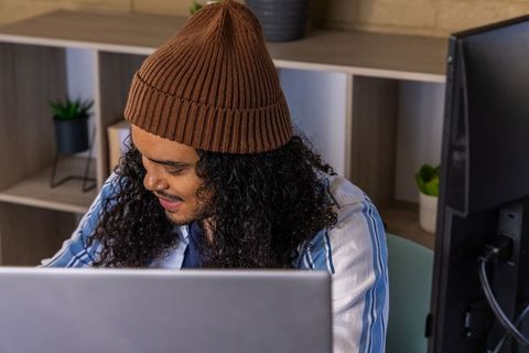 Young man wearing brown beanie typing on laptop in modern home office workspace
