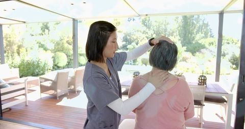 Caregiver assessing senior neck mobility on sunlit patio for home elder care therapy