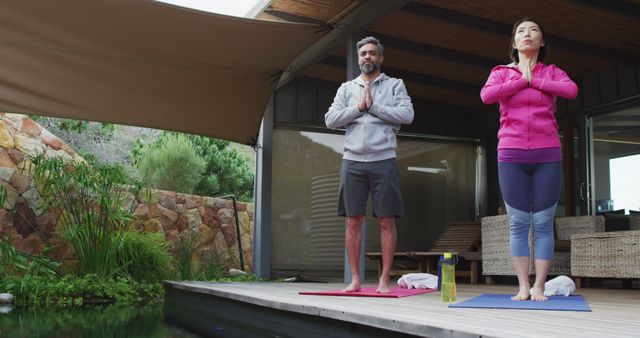 Couple engaging in yoga practice on comfortable patio near tranquil koi pond. Both wear athletic clothing and stand in prayer pose, demonstrating mindfulness and concentration. Suitable for concepts of wellness, relaxation, nature, and couple bonding. Ideal for promoting outdoor yoga classes, fitness retreats, mindfulness practices, and healthy living.