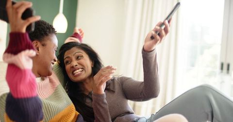 Joyful female friends bonding on sofa with smartphones