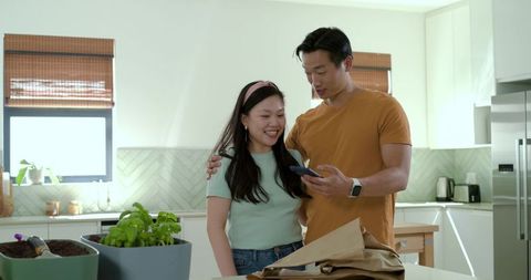 Couple checking smartphone while unpacking groceries and herbs on bright kitchen island