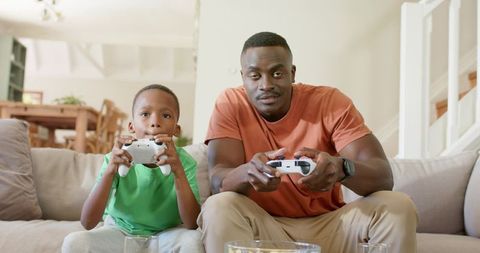 African american father and son playing video games together on bright living room sofa