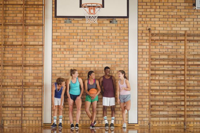 Group of high school kids standing and chatting in a basketball court. They are smiling and appear to be enjoying their time together. This image can be used for promoting youth sports, teamwork, school activities, and friendship. It is ideal for educational materials, sports programs, and community events.
