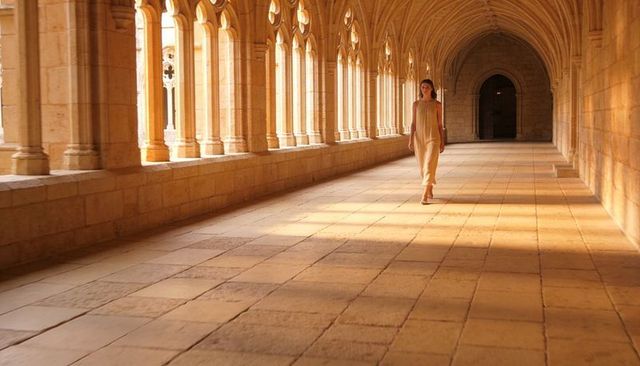 Woman walking through sunlit gothic cloister with dramatic arches and long perspective