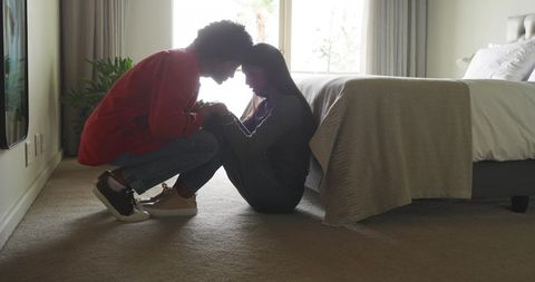 Diverse couple sharing intimate moment on bedroom floor, leaning foreheads, holding hands