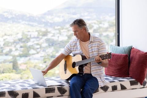 Mature man practicing acoustic guitar on window bench while using laptop with scenic view