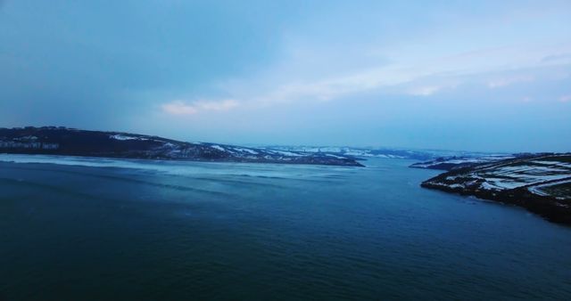 A breathtaking aerial view shows a coastal landscape covered in snow during dusk. The scene showcases the natural beauty of the shoreline under a cloudy sky, creating a serene and peaceful atmosphere. This image is ideal for use in travel brochures, nature documentaries, winter-themed promotions, and scenic background content. It can also be used to evoke a sense of tranquility and natural wonder in various design projects.