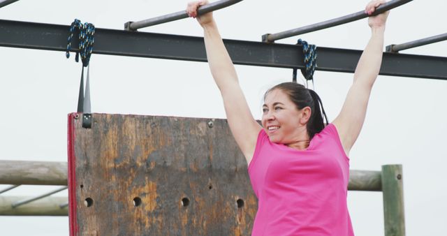 Smiling caucasian woman in pink t shirt hanging from monkey bars on bootcamp training course. Female fitness, challenge and healthy lifestyle.