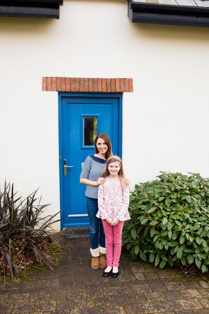 Mother and daughter standing near house entrance with blue door, smiling and bonding. Ideal for use in family-oriented advertisements, real estate promotions, home improvement campaigns, and lifestyle blogs.