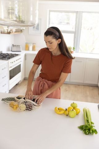 Woman arranging groceries into reusable mesh bag on bright modern kitchen island