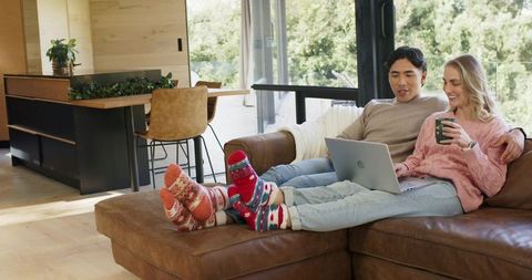 Relaxing mixed-race couple on brown leather sectional using laptop and sharing coffee