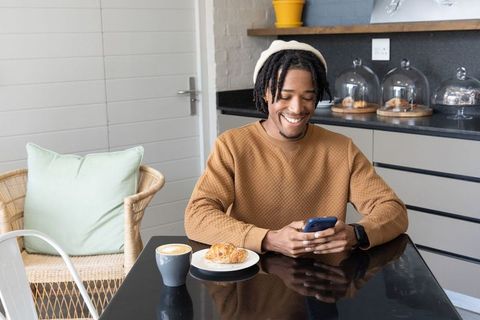 African american man smiling and checking smartphone in cafe with latte and croissant