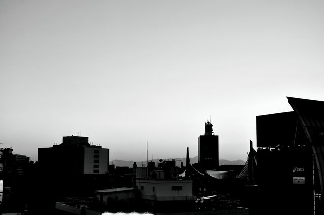 This black and white image captures a city skyline at dusk, presenting a moody and dramatic atmosphere. The buildings form dark silhouettes against the light sky, providing high contrast and highlighting architectural shapes. Perfect for projects focused on urban life, architecture, or atmospheric visuals. Can be used in creative designs, urban studies, and industrial themes.