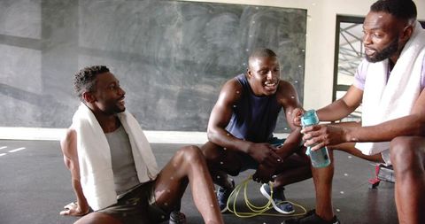 Three men resting and sharing water bottle after intense gym workout