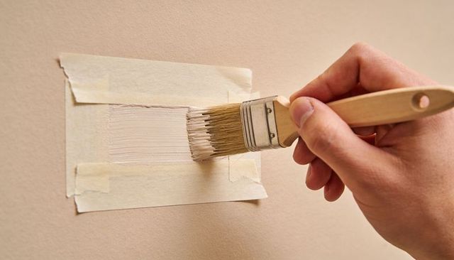 Right Hand Holding Paintbrush Applying Beige Paint Sample on Wall with Masking Tape
