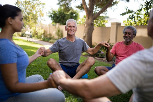 Group of seniors meditating with a young trainer in a park, holding hands and smiling. Ideal for promoting wellness, fitness programs, mindfulness practices, and intergenerational activities. Perfect for use in health and wellness campaigns, community event promotions, and lifestyle blogs.