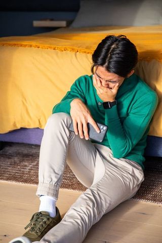 Young man sitting on floor holding smartphone and covering mouth, anxious bedroom moment
