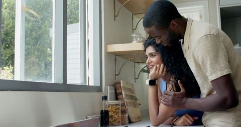 Sunlit kitchen couple embracing by window, leaning on countertop, intimate lifestyle moment