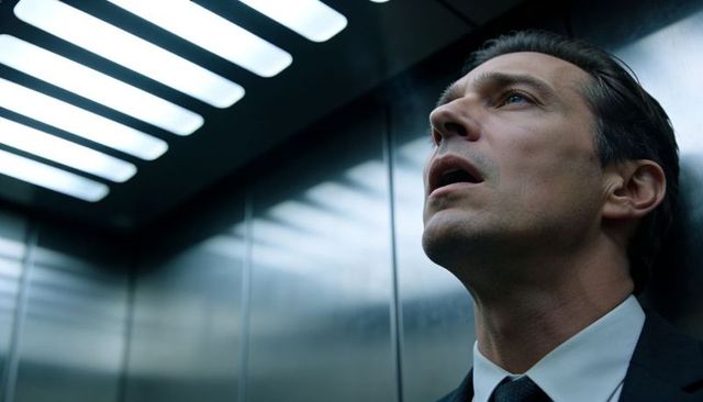 Businessman craning neck in stainless elevator under fluorescent lights conveying tension