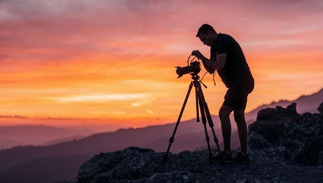 Silhouette photographer adjusting telephoto camera on mountain ridge at vibrant sunset