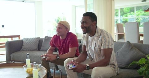 Friends playing video games on gray sofa with snacks and drinks in bright open living room
