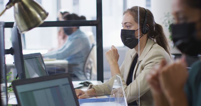 Caucasian businesswoman wearing face mask at desk with sneeze shield talking wearing phone headset. independent creative business in a modern office during coronavirus covid 19 pandemic.
